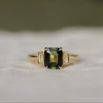 Gold ring with a green rectangular gemstone set with claws with 2 bezel set baguette diamonds on a white fabric background
