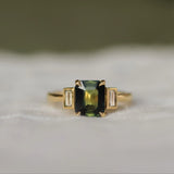 Gold ring with a green rectangular gemstone set with claws with 2 bezel set baguette diamonds on a white fabric background