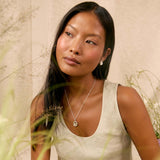 A woman wearing a chunky silver knot pendant necklace, styled with a beige linen tank top and skirt against a neutral textured wall with dried grass.