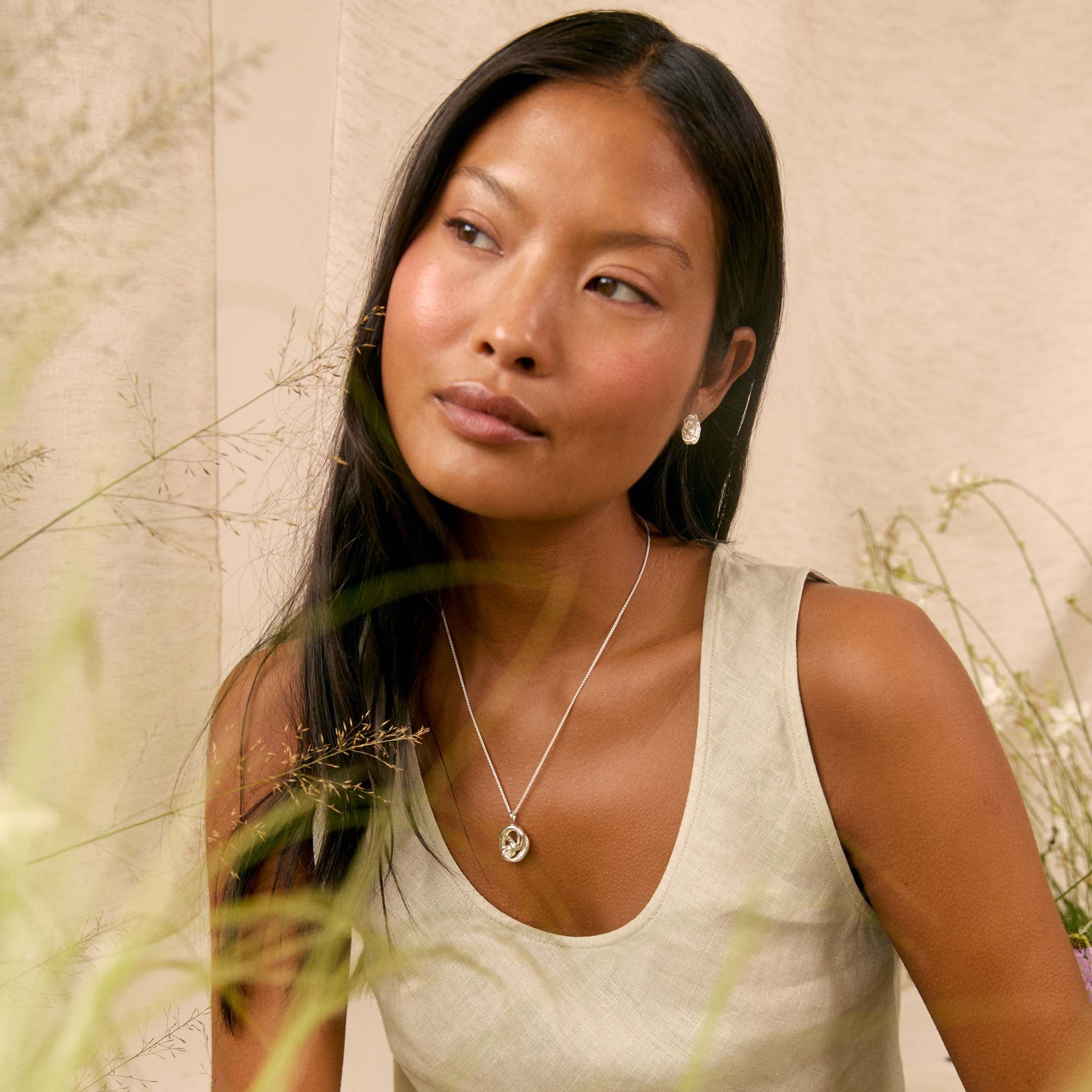 A woman wearing a chunky silver knot pendant necklace, styled with a beige linen tank top and skirt against a neutral textured wall with dried grass.