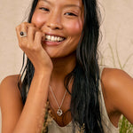 A smiling woman with long dark hair models a silver pendant necklace with a knot design, a silver ring, and a silver bangle against a warm beige background with dried grass.