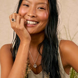 A smiling woman with long dark hair models a silver pendant necklace with a knot design, a silver ring, and a silver bangle against a warm beige background with dried grass.