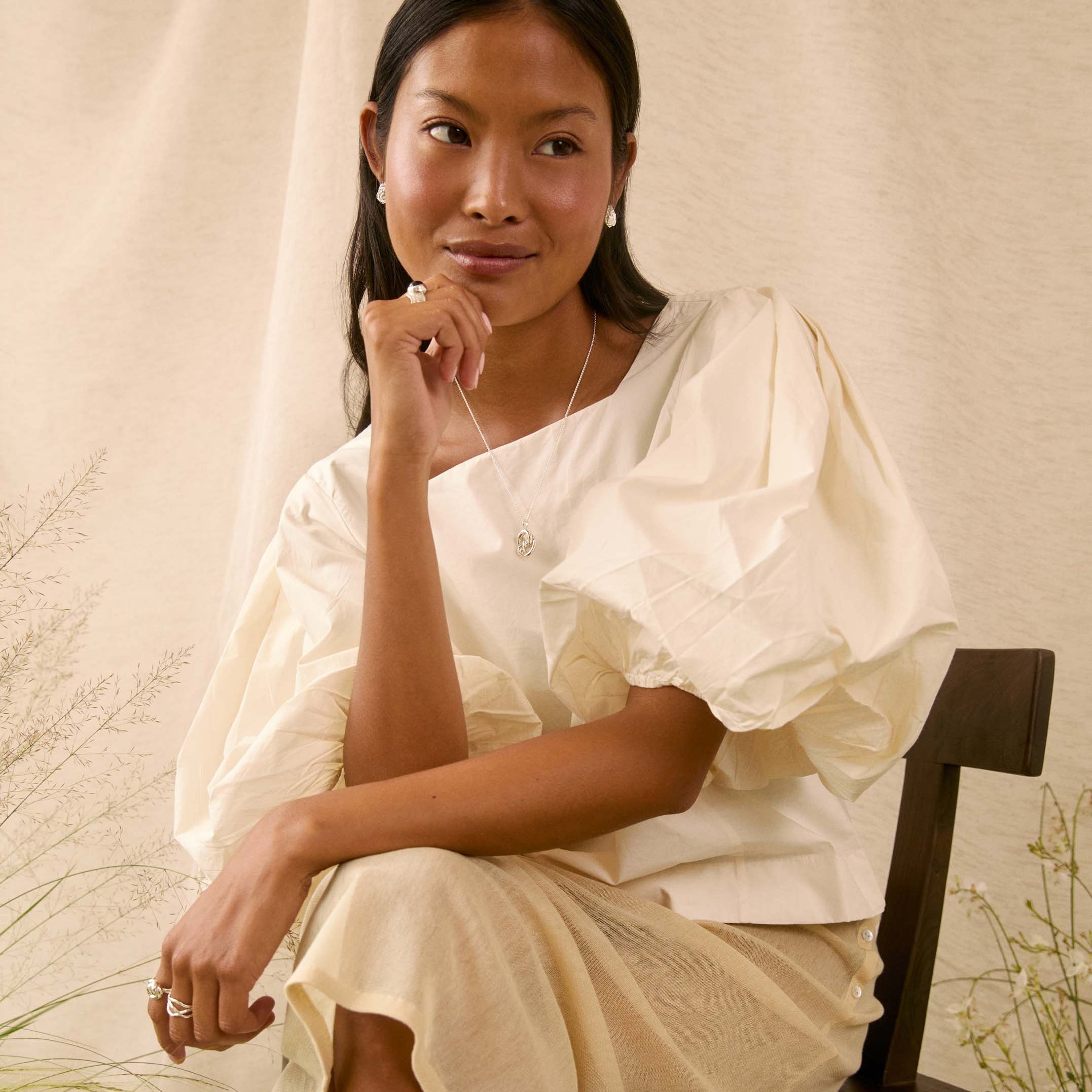 A woman sits on a dark wooden chair wearing a cream-colored, short puff-sleeve dress with a draped neckline. The background consists of a draped beige fabric backdrop with some dried botanical elements on the sides.