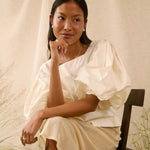 A woman sits on a dark wooden chair wearing a cream-colored, short puff-sleeve dress with a draped neckline. The background consists of a draped beige fabric backdrop with some dried botanical elements on the sides.