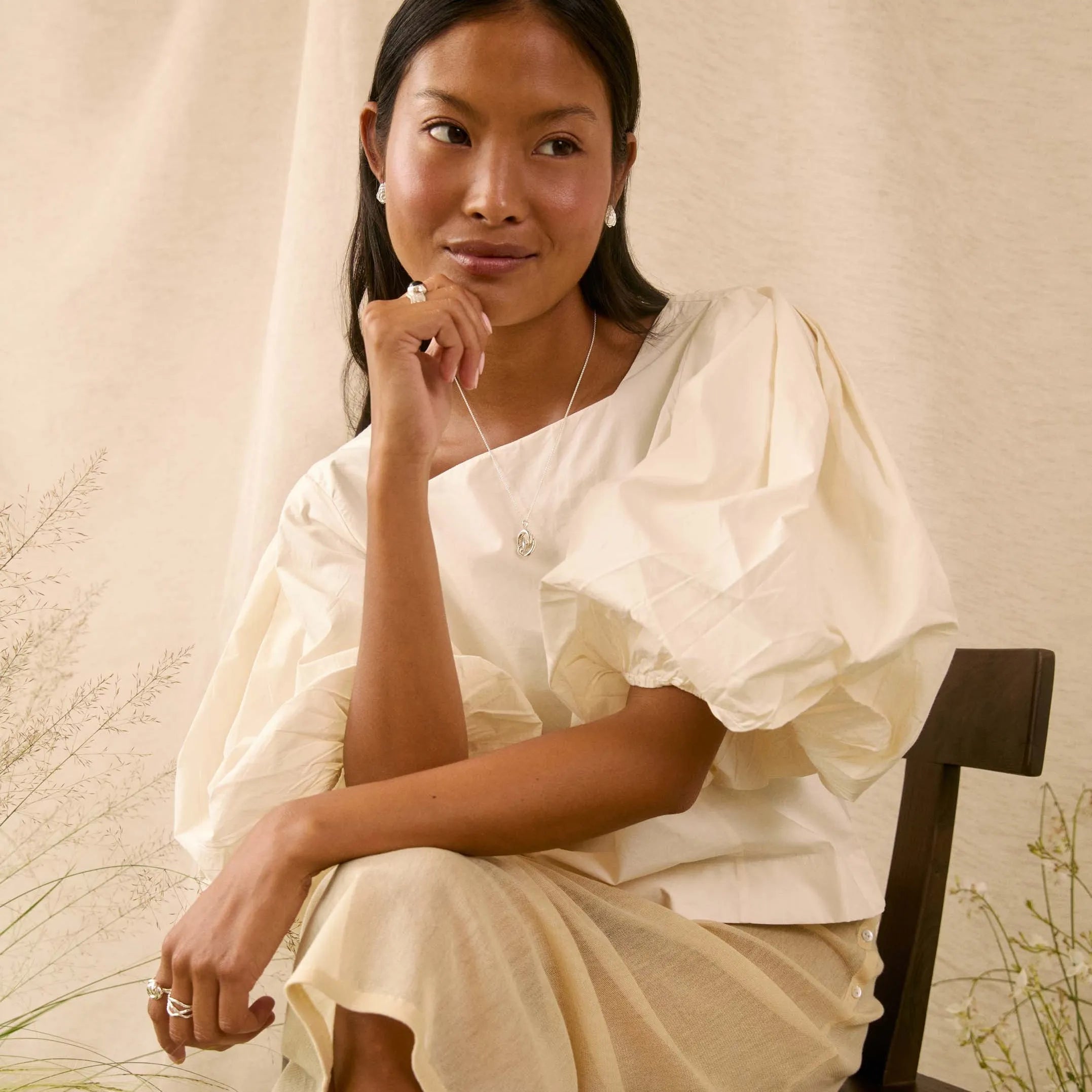 A woman sits on a dark wooden chair wearing a cream-colored, short puff-sleeve dress with a draped neckline. The background consists of a draped beige fabric backdrop with some dried botanical elements on the sides.