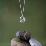 A silver pendant necklace with an abstract interlocking knot design hangs above a small stack of smooth river stones on a dark olive green background.
