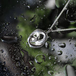 A silver pendant necklace with a polished, interlocking knot design, resting on a wet glass surface covered in water droplets.