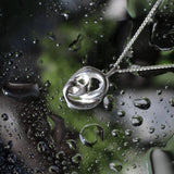 A silver pendant necklace with a polished, interlocking knot design, resting on a wet glass surface covered in water droplets.