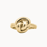 A gold ring featuring a sculpted, textured knot design on a smooth band, shown against a plain off-white background.