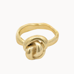 A gold ring featuring a sculptural, organic knot design on a plain light background.