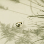 A delicate gold ring with a knot design, photographed on a warm beige surface with soft, artistic shadows of dried grasses.