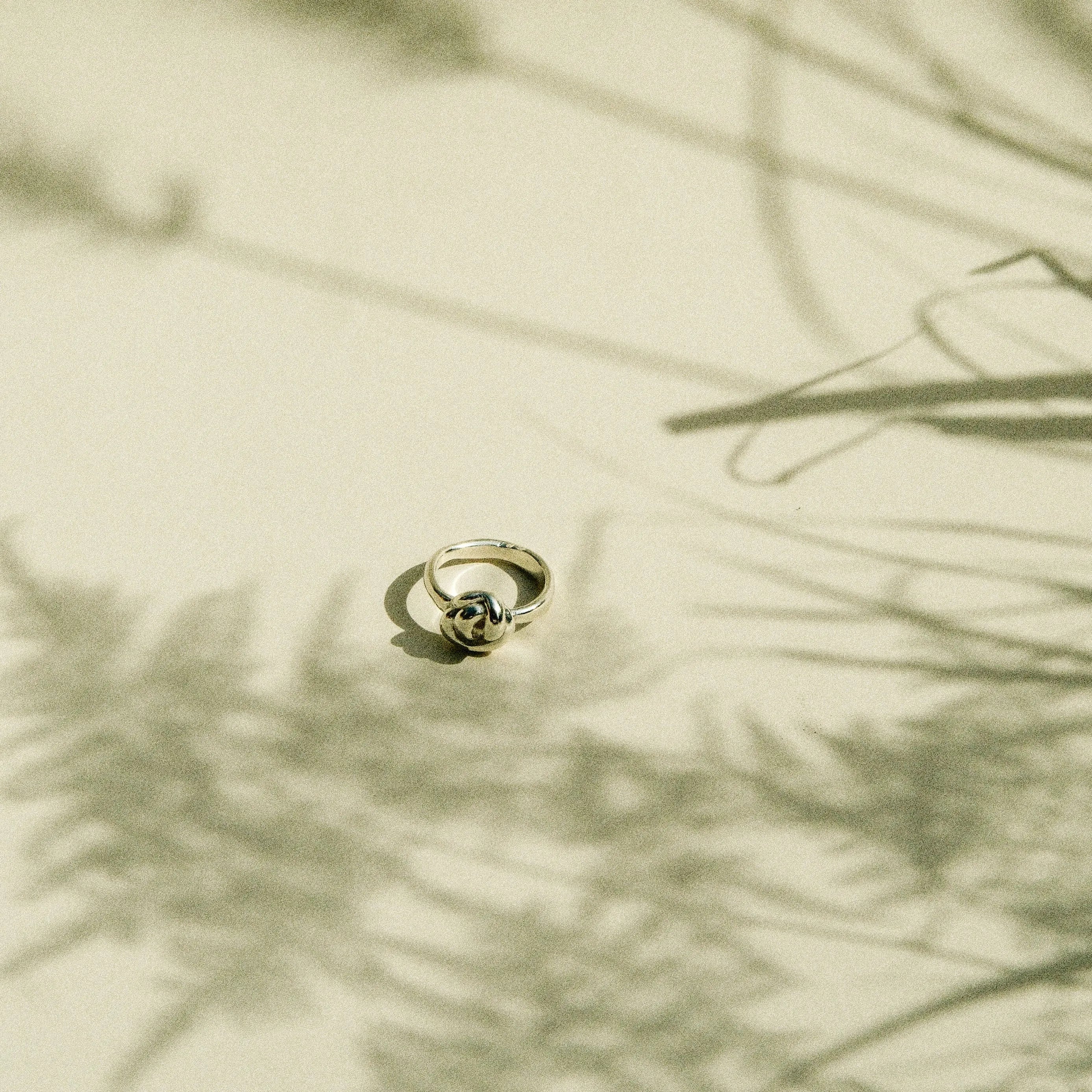 A delicate gold ring with a knot design, photographed on a warm beige surface with soft, artistic shadows of dried grasses.