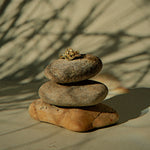 A small gold ring is stacked on top of three balanced stones against a warm beige background with soft shadows of tree branches.
