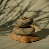 A small gold ring is stacked on top of three balanced stones against a warm beige background with soft shadows of tree branches.