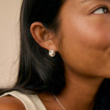 A close-up lifestyle shot of a woman wearing a small silver twisted knot stud earring and a silver chain necklace with a silver pendant, set against a warm beige background.
