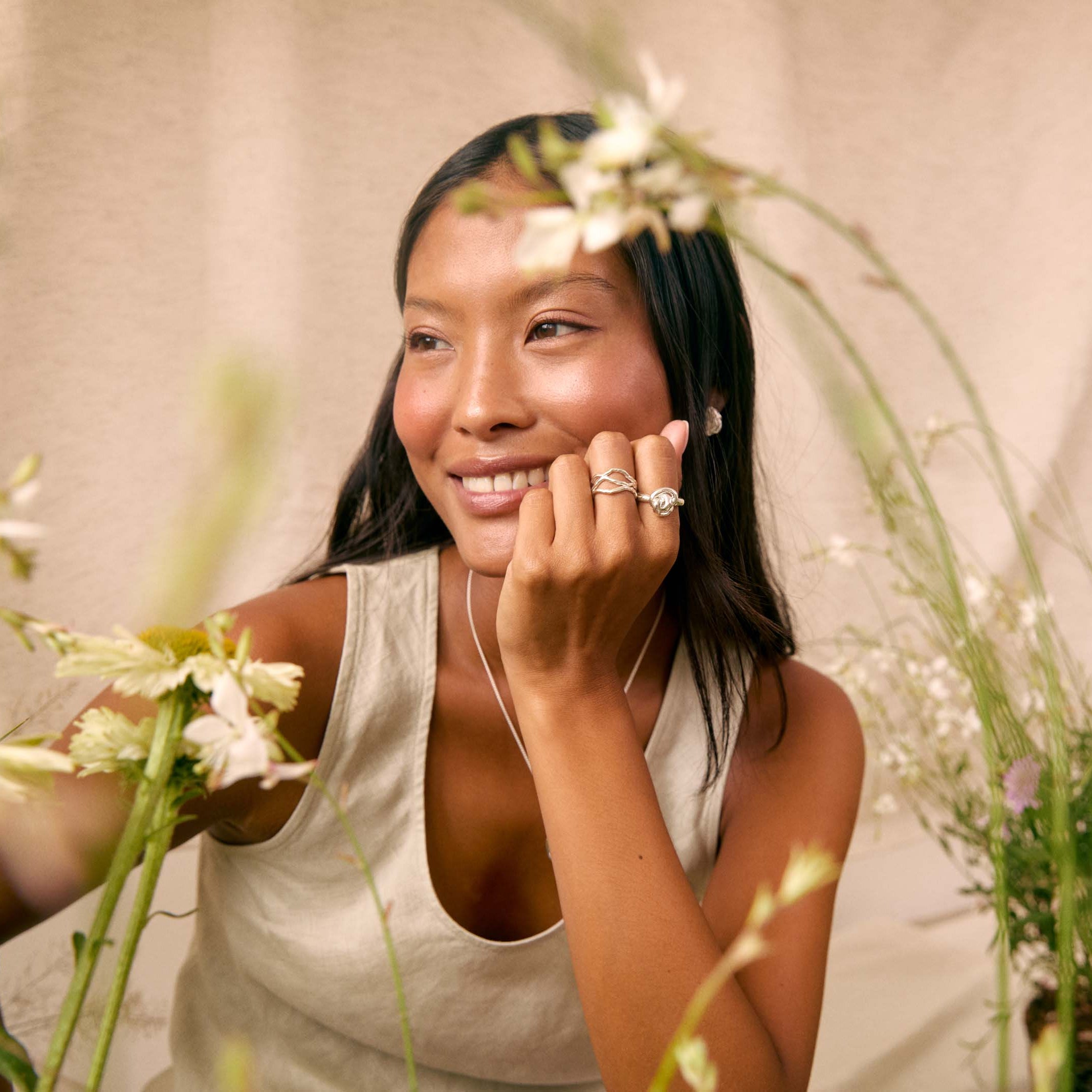 A woman with long dark hair smiles while wearing delicate silver rings on her fingers, posed among soft white flowers and green foliage against a warm beige backdrop.