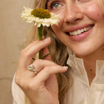 A smiling blonde woman in a cream blouse holds a daisy flower near her face, wearing a silver ring on one finger and a small earring in her other ear. The background is a soft beige wall with subtle dried floral decor.