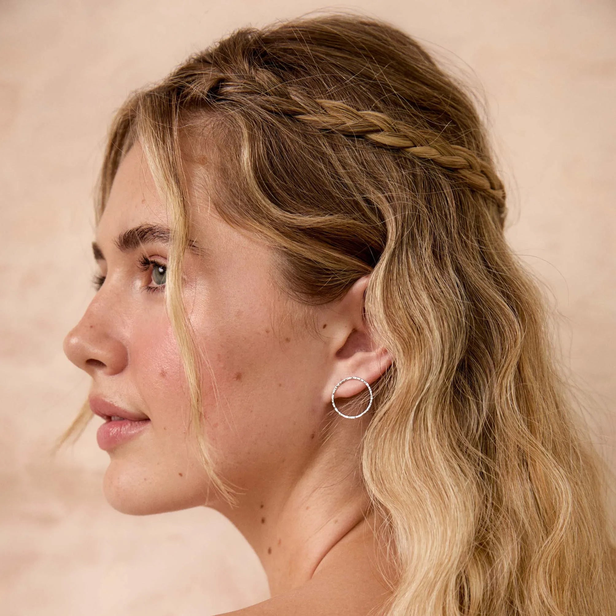 A woman shown in side profile wearing a delicate silver hoop earring, with long blonde wavy hair and a braided detail across the top of her head, against a warm beige background.