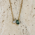 A delicate gold chain necklace with a unique cluster pendant featuring a green oval-cut green sapphire, a pear diamond and small round diamond, displayed on a textured beige stone surface.