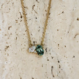 A delicate gold chain necklace with a unique cluster pendant featuring a green oval-cut green sapphire, a pear diamond and small round diamond, displayed on a textured beige stone surface.