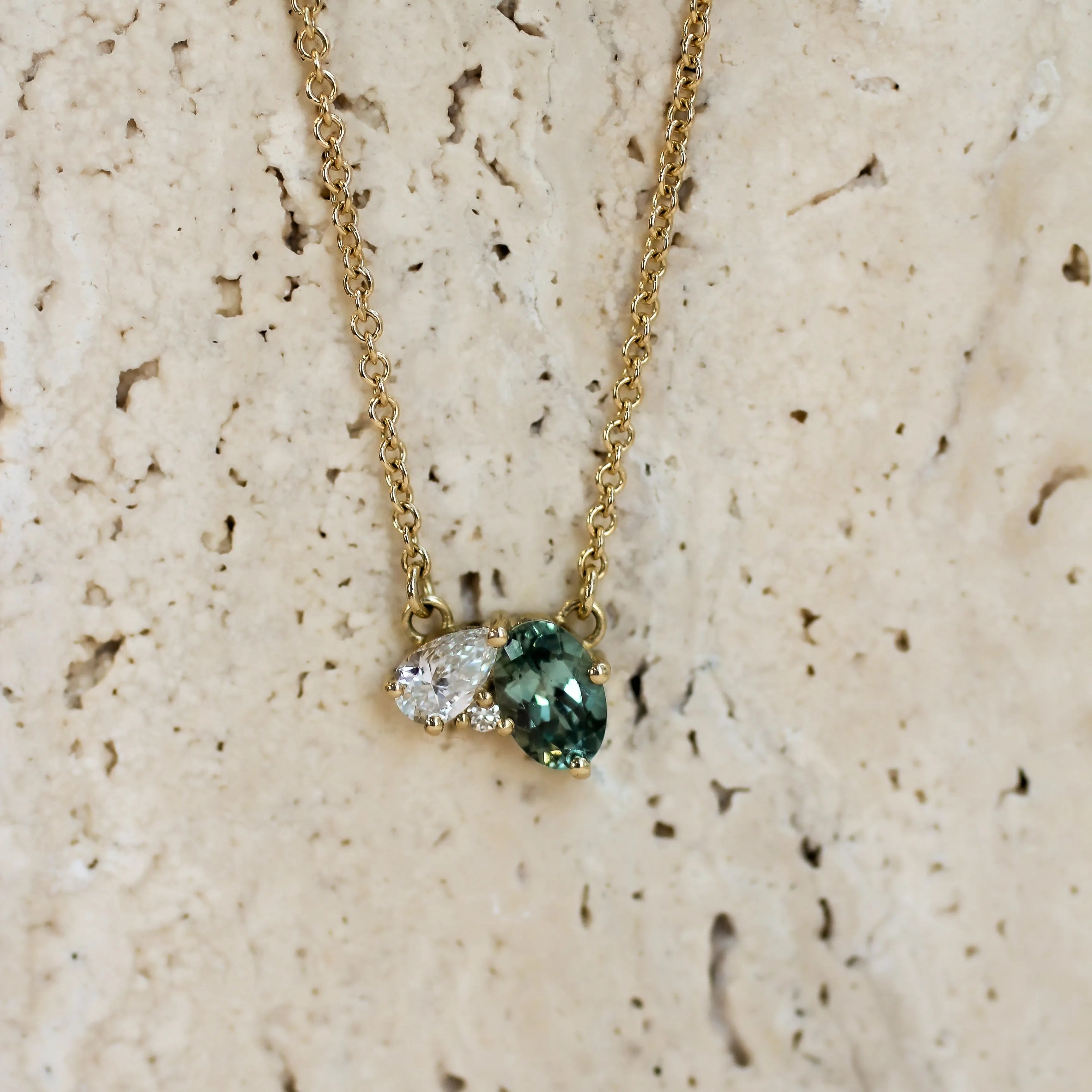 A delicate gold chain necklace with a unique cluster pendant featuring a green oval-cut green sapphire, a pear diamond and small round diamond, displayed on a textured beige stone surface.