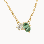 A gold chain necklace with an greeny blue sapphire pendant alongside a marquise-cut diamond, shown on a plain white background.