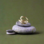 A gold ring with a sculptural, flowing s-shaped design, displayed on a stack of smooth gray zen stones against a solid olive green background.