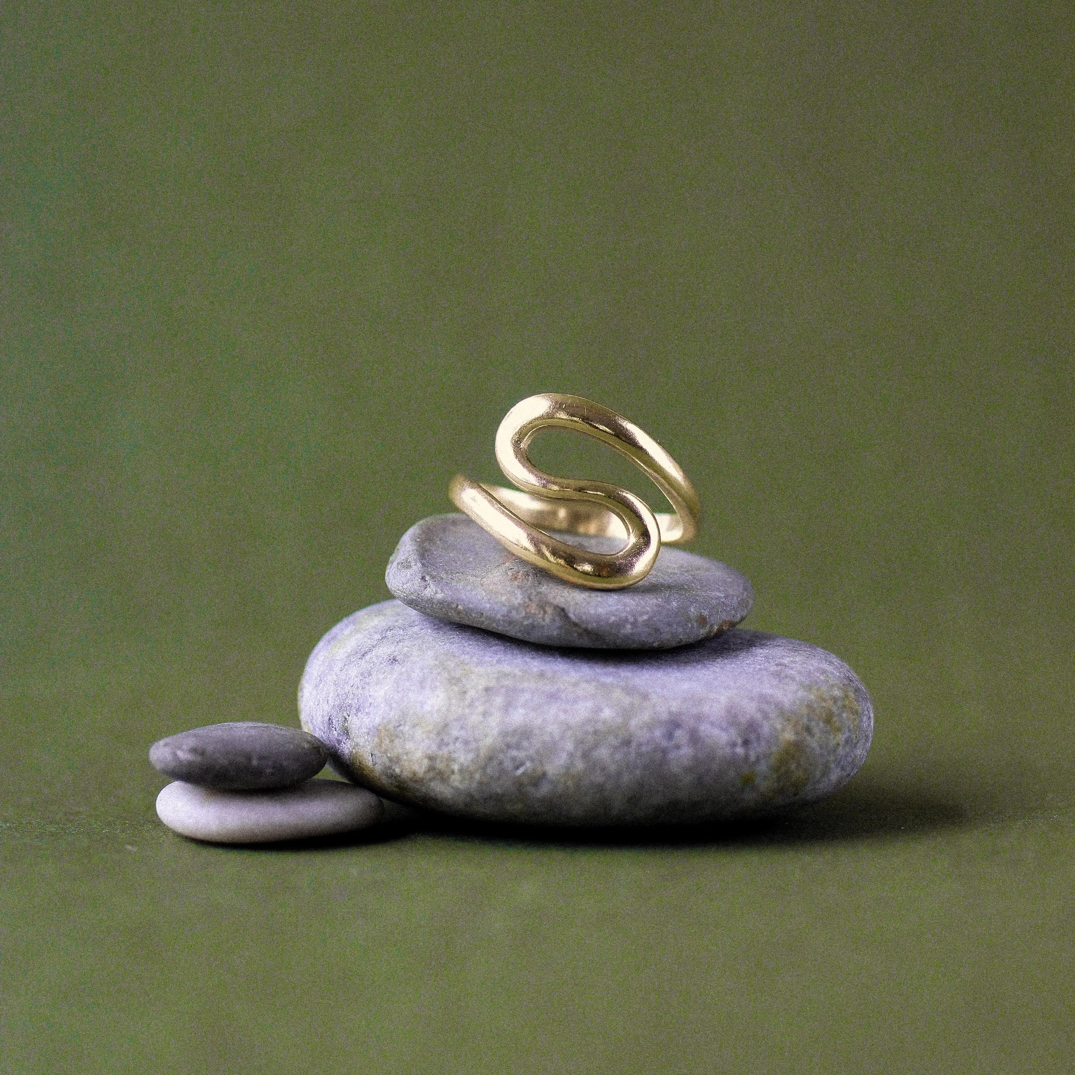A gold ring with a sculptural, flowing s-shaped design, displayed on a stack of smooth gray zen stones against a solid olive green background.