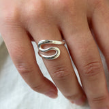 A close-up of a hand wearing a polished silver ring with a stylized, flowing s-shaped design, resting on a textured white fabric background.