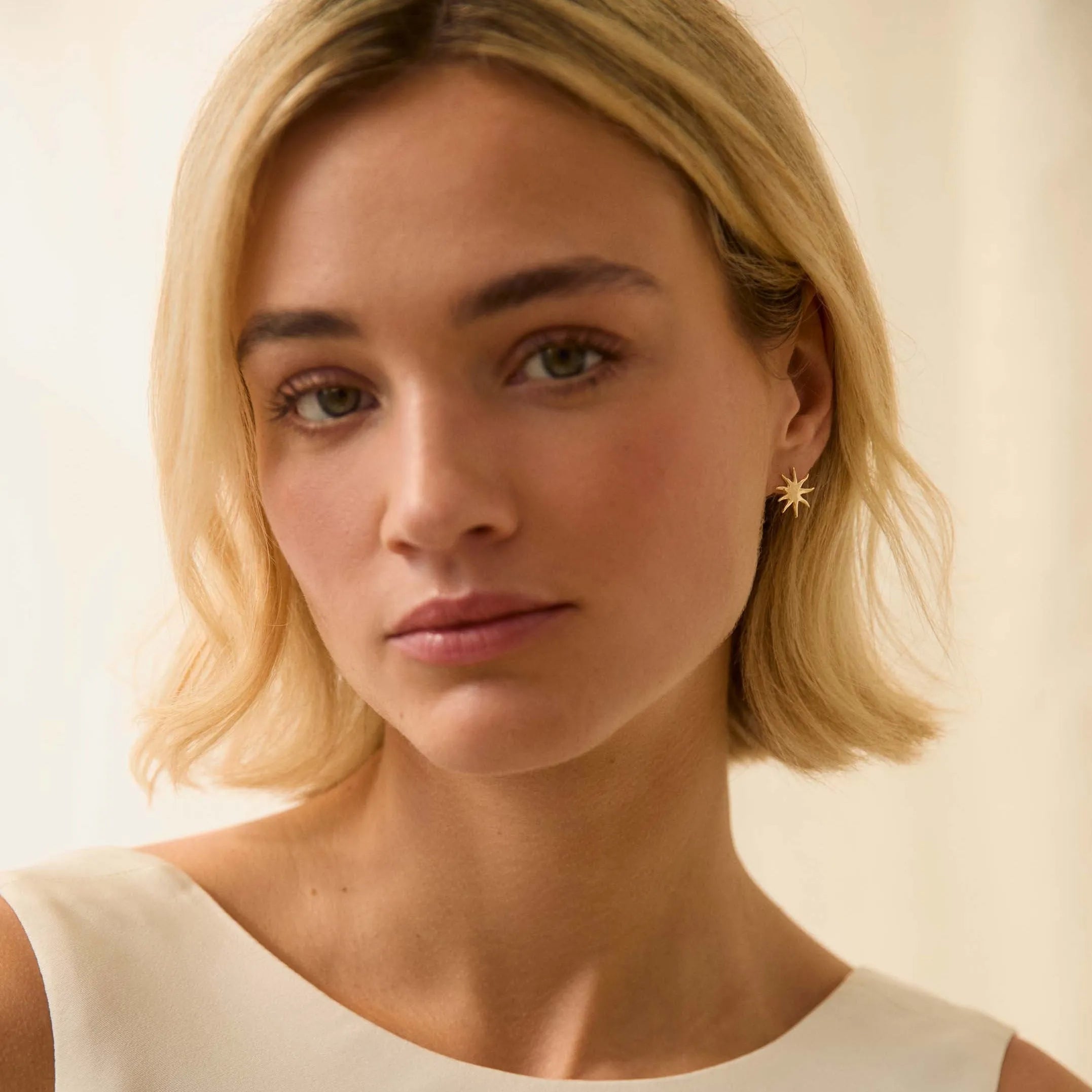 Woman with blonde hair wearing gold sun stud earrings and a white top against a beige background