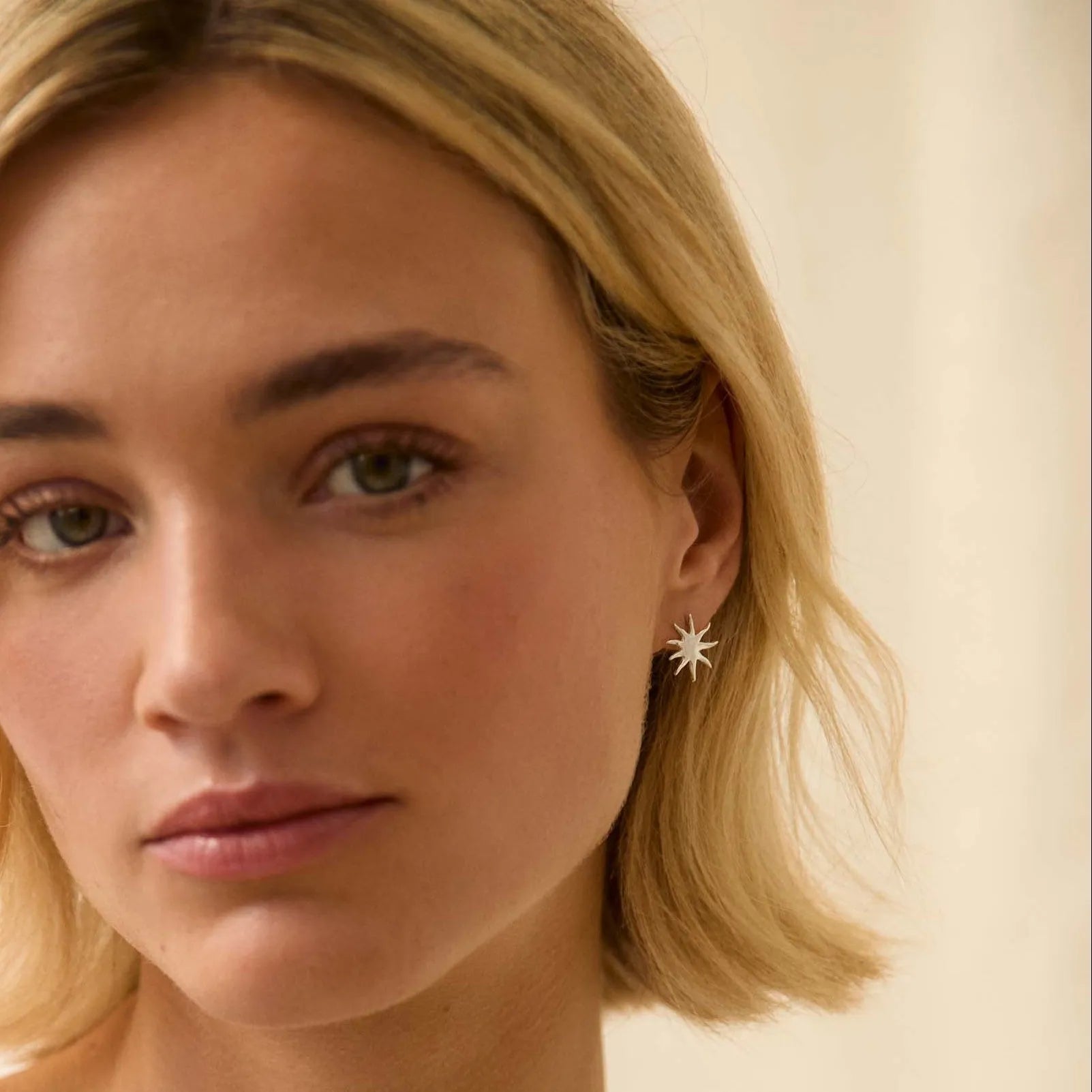 Woman with blonde hair wearing silver sun shaped stud earrings against a beige background