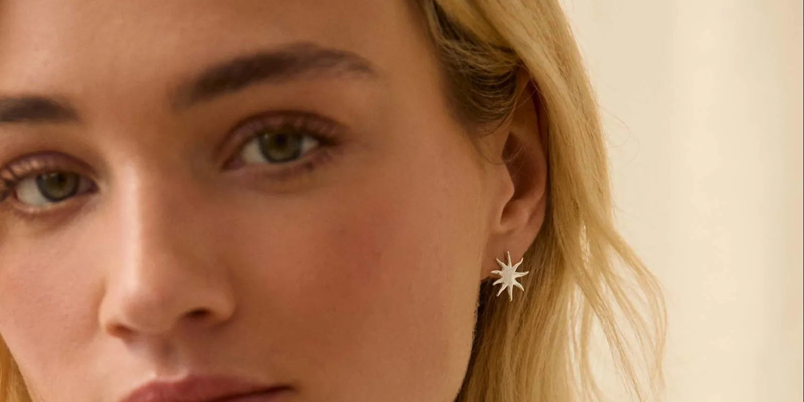 Woman with blonde hair wearing silver sun shaped stud earrings against a beige background