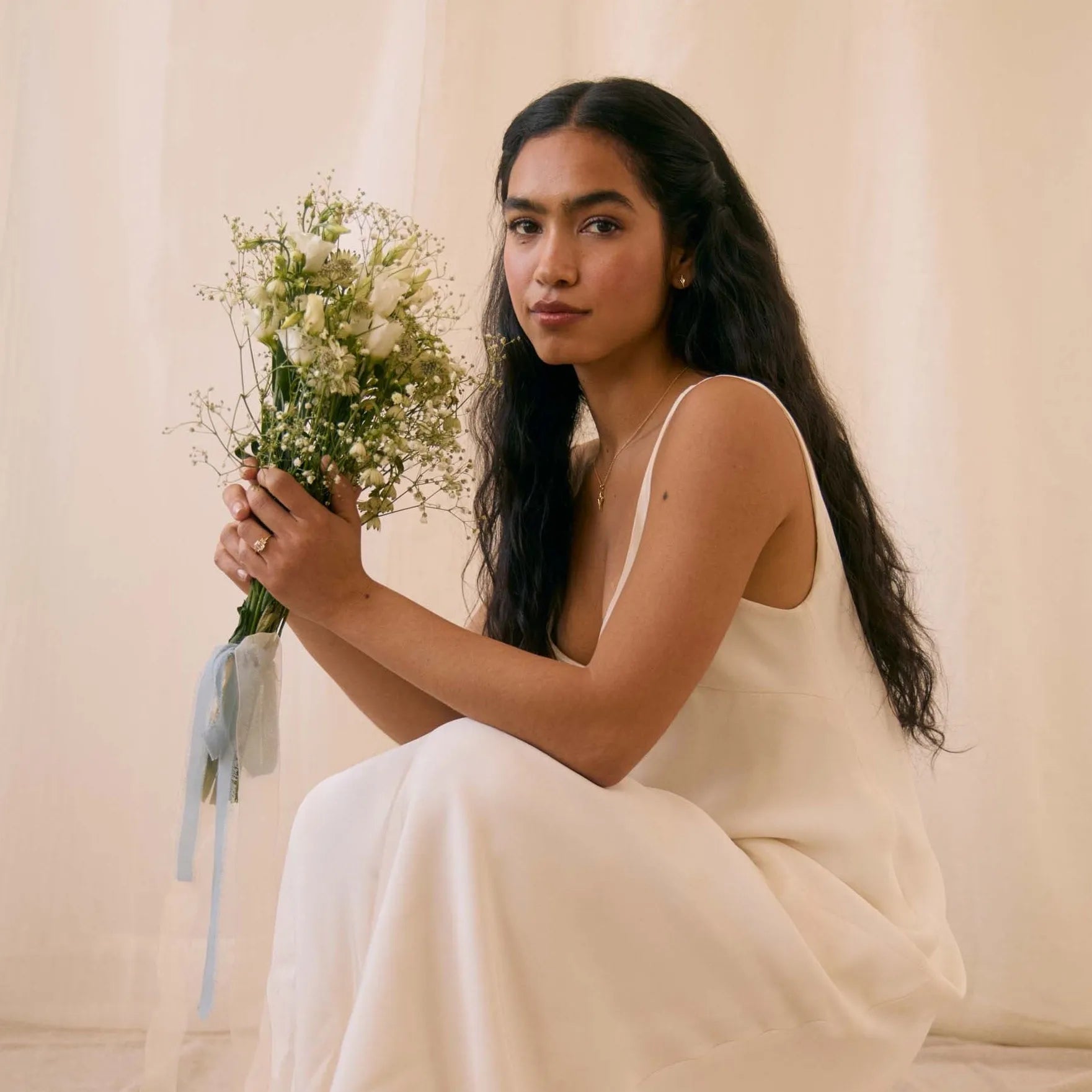 Woman in a white dress wearing tiny star stud earrings, holding a bouquet of flowers against a neutral background