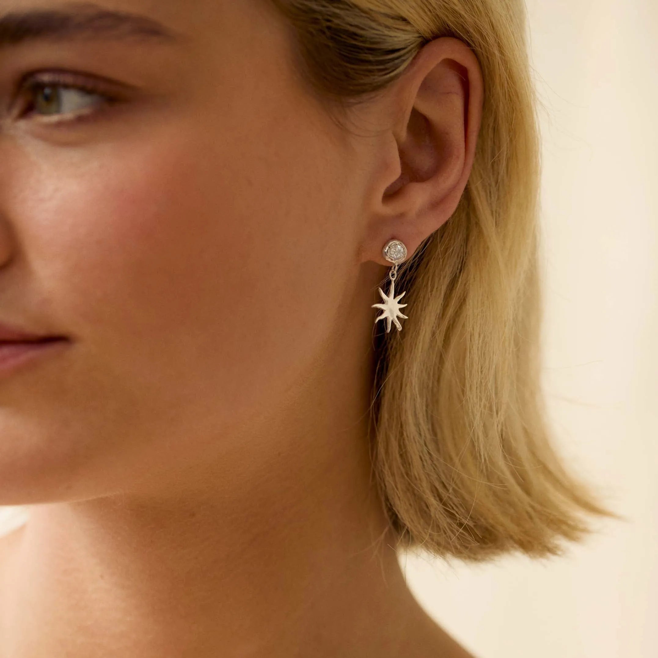 Close-up of a blonde woman wearing diamond studs with sun-shaped dangle charms with a neutral background
