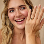 A smiling blonde woman wearing a gold ring with a small green stone and smaller diamonds, holding up her hand to show the ring. She is wearing a white ruffled top against a soft beige background.