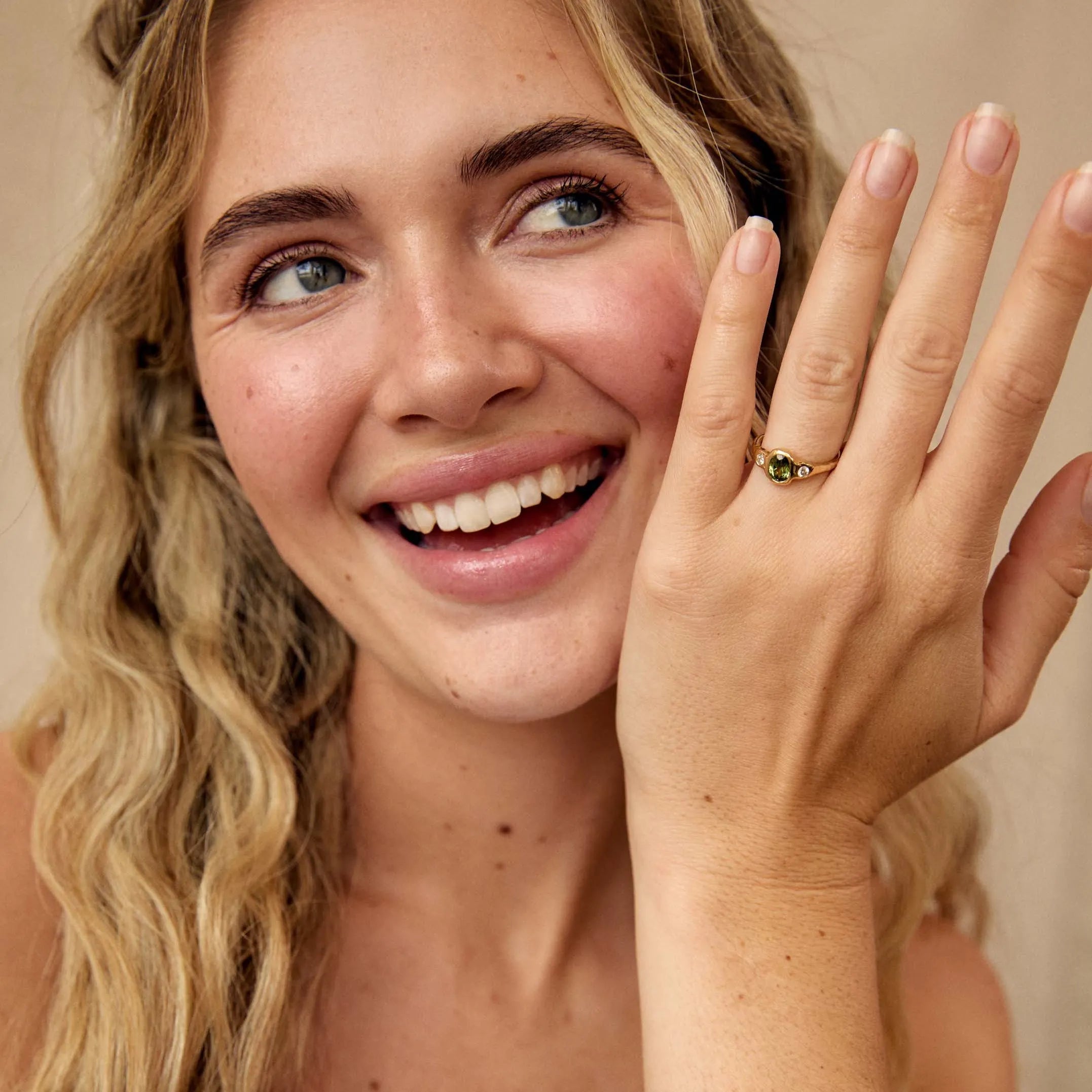 A smiling blonde woman wearing a gold ring with a small green stone and smaller diamonds, holding up her hand to show the ring. She is wearing a white ruffled top against a soft beige background.