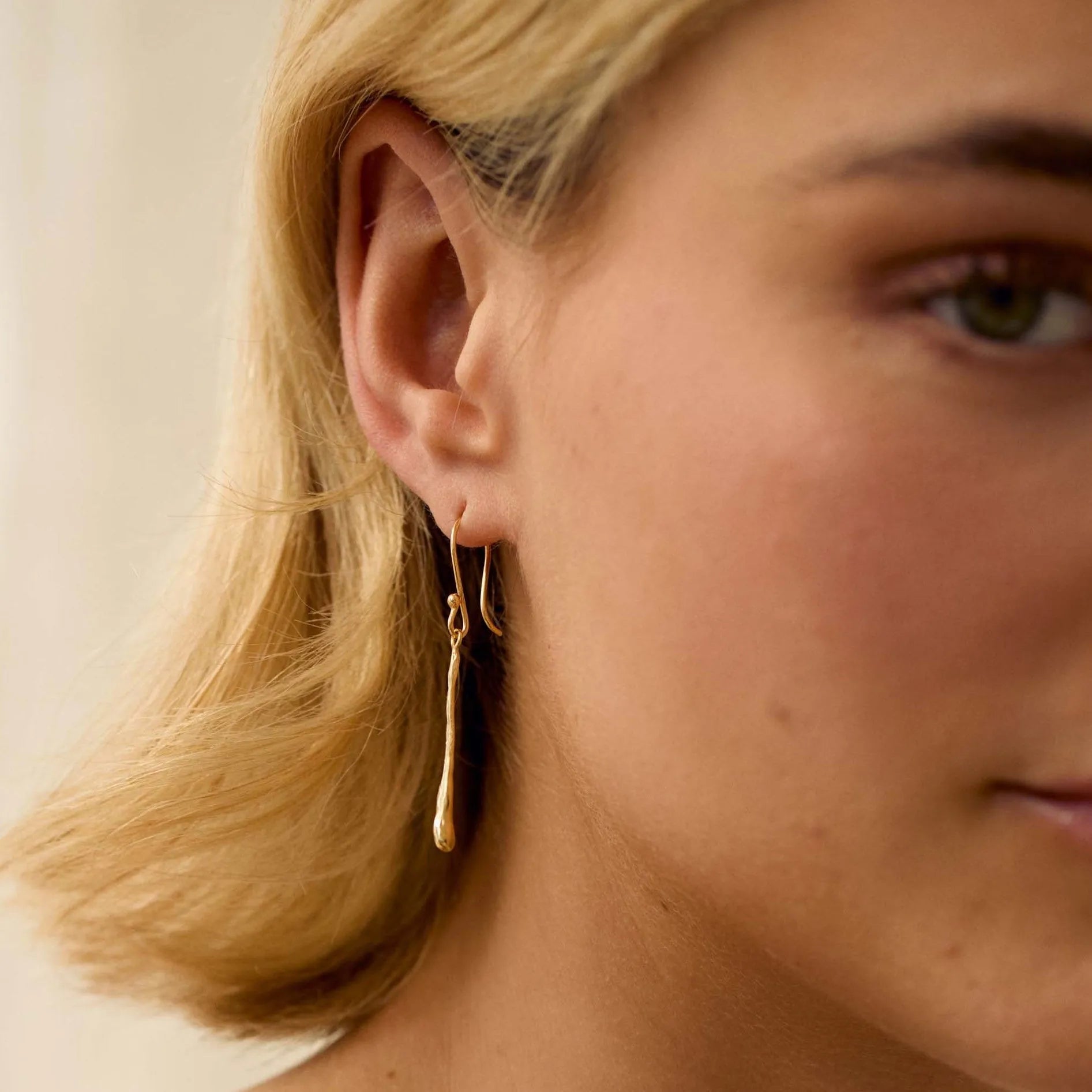 Close-up of a woman wearing gold dangly earrings with molten gold bar detail against a neutral background