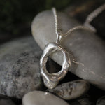 A silver abstract organic-shaped pendant necklace resting on smooth gray river stones with a dark, blurred natural background.