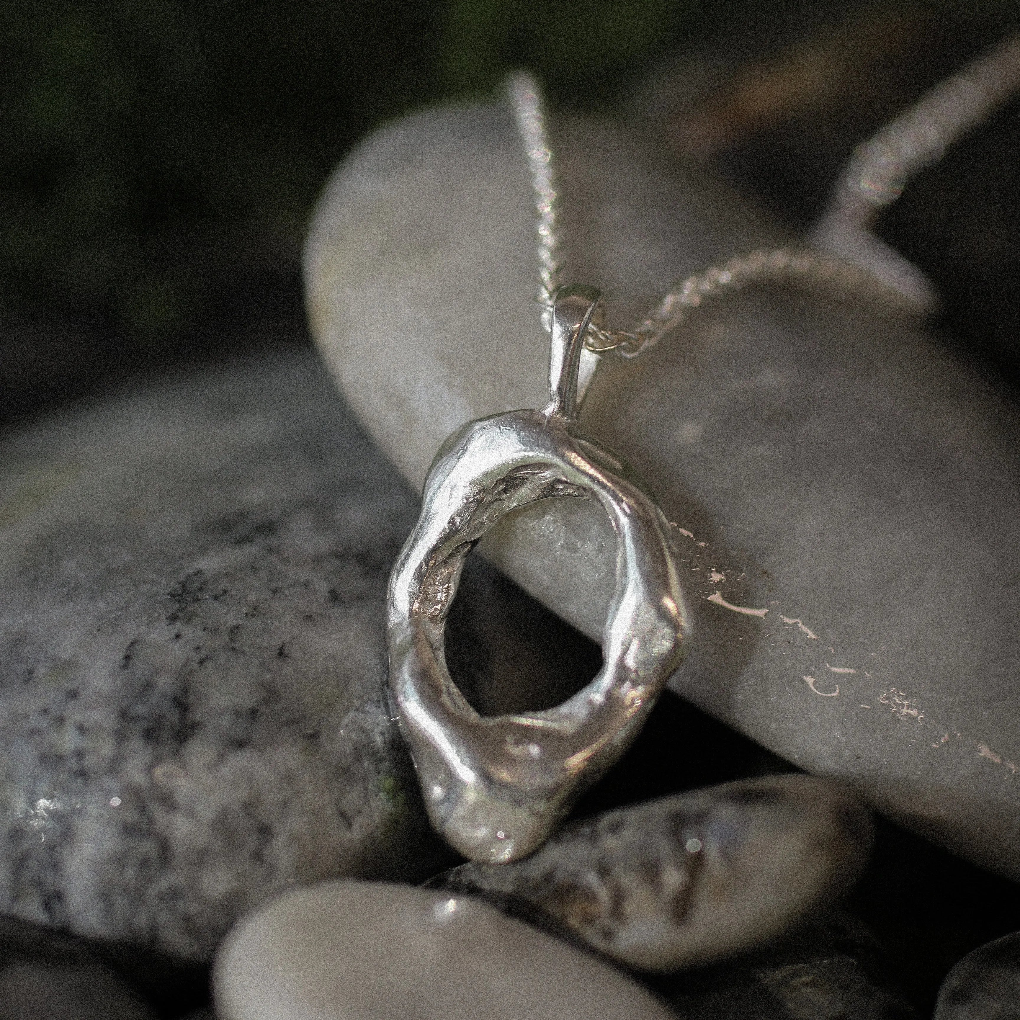 A silver abstract organic-shaped pendant necklace resting on smooth gray river stones with a dark, blurred natural background.