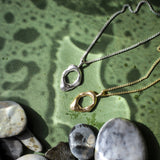 Two abstract open pendant necklaces, one silver-toned and one gold-toned, displayed on a textured green surface with smooth river stones.