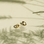 Two small gold hoop-style earrings resting on a warm beige surface with soft shadows cast across the scene.