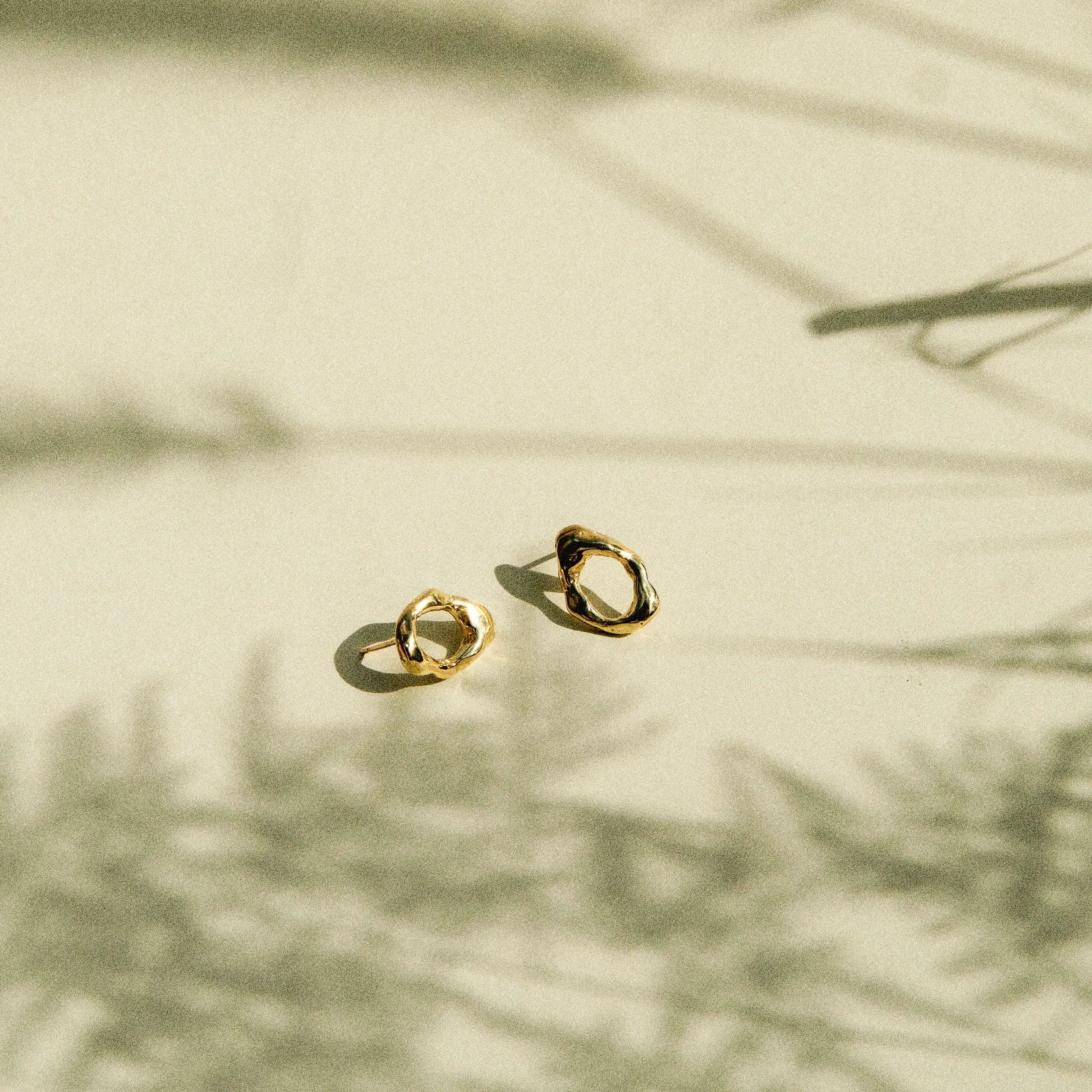 Two small gold hoop-style earrings resting on a warm beige surface with soft shadows cast across the scene.