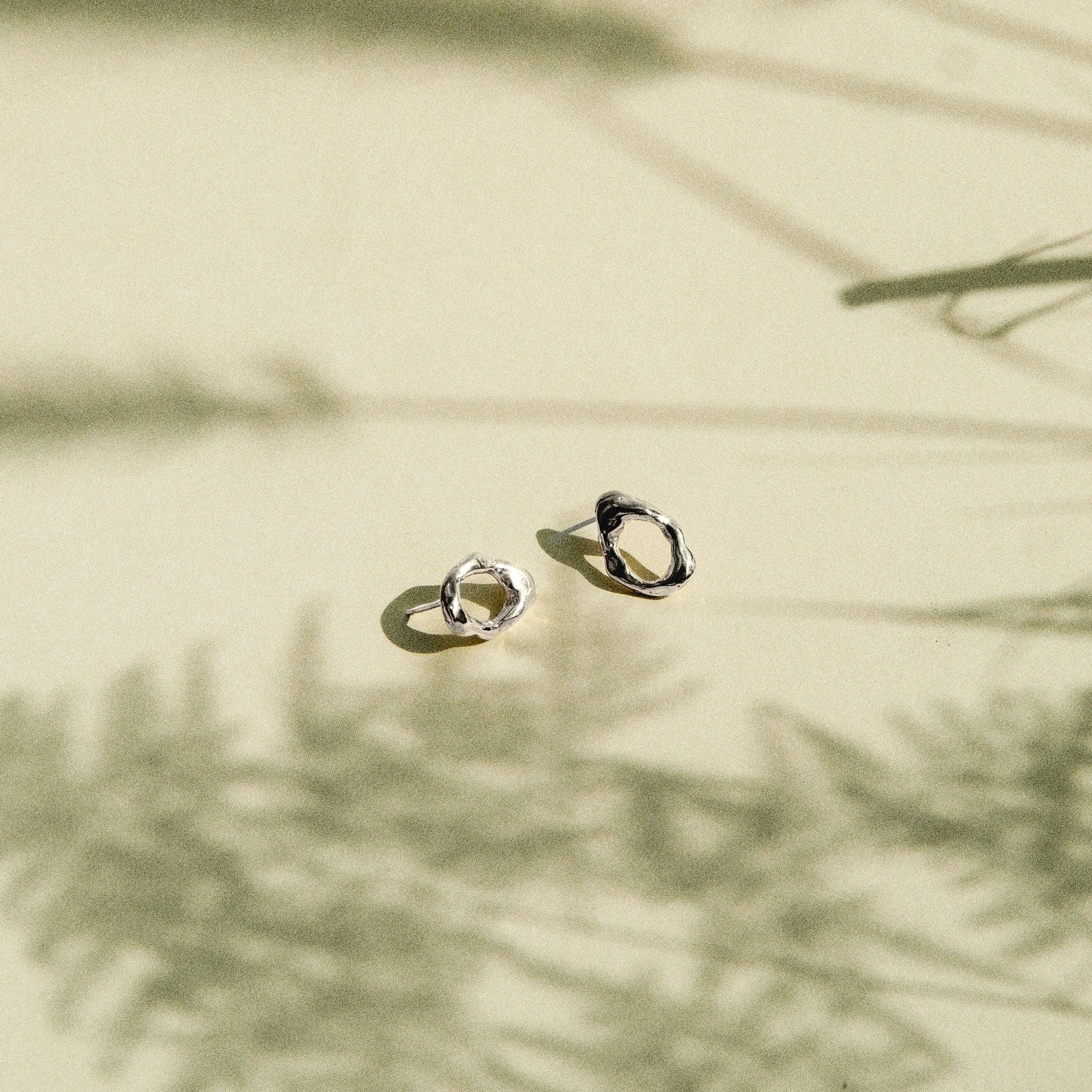 Two small gold hoop-style earrings resting on a warm beige surface with soft shadows cast across the scene.