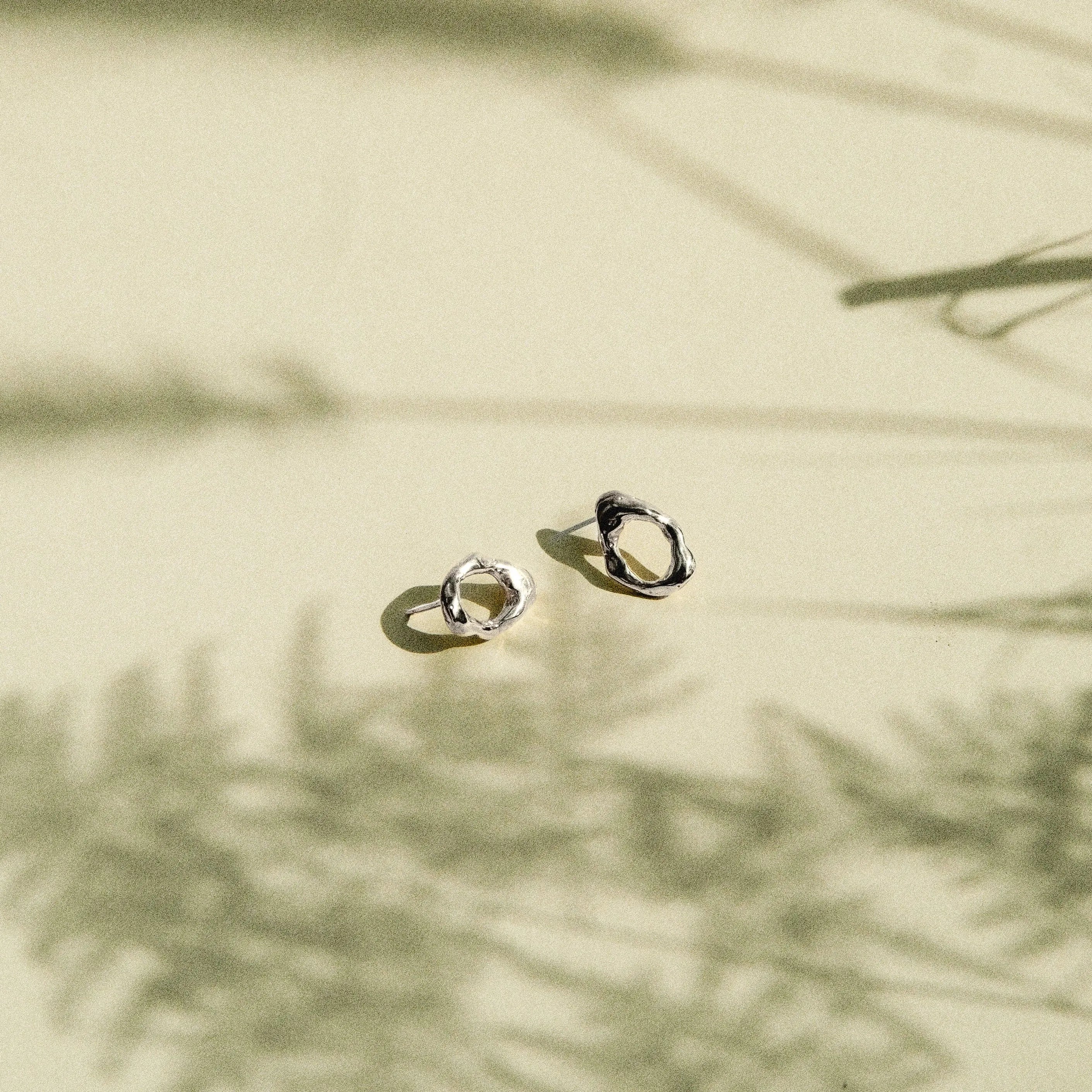 Two small gold hoop-style earrings resting on a warm beige surface with soft shadows cast across the scene.