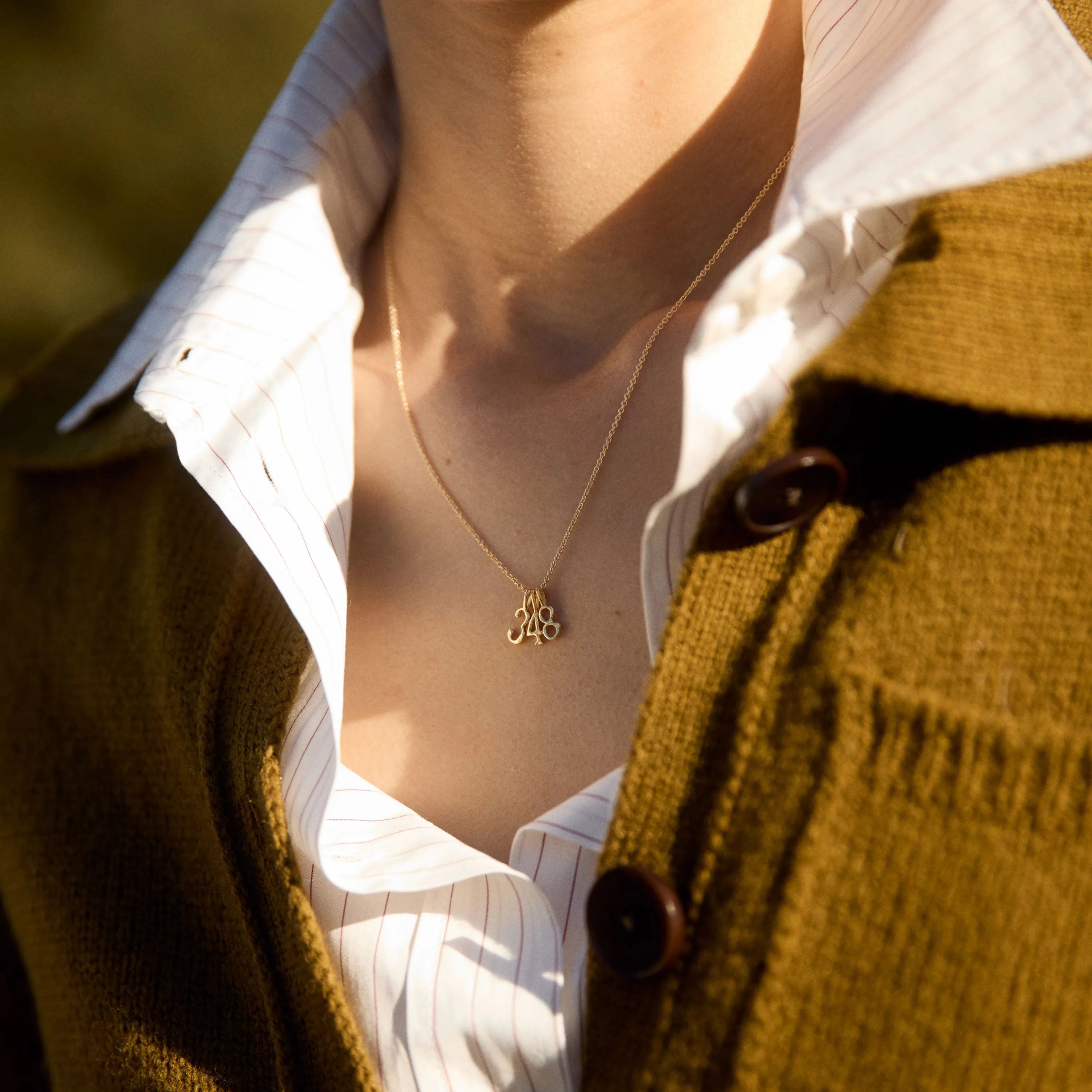 Person wearing a mustard-colored cardigan over a white shirt with a necklace featuring a three number pendants.