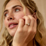 A close-up lifestyle shot of a woman wearing a gold diamond trilogy engagement ring and textured rustic wedding ring looking upward with a soft, dreamy expression against a warm beige background.