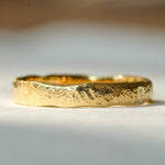 A textured, organic-shaped gold ring resting on a light gray surface with a soft, blurred background.