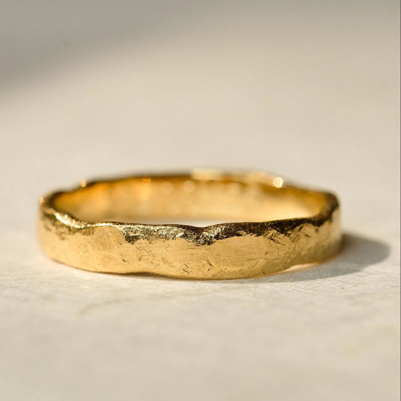 A textured, hammered gold band ring resting on a neutral, light beige surface with soft shadows.