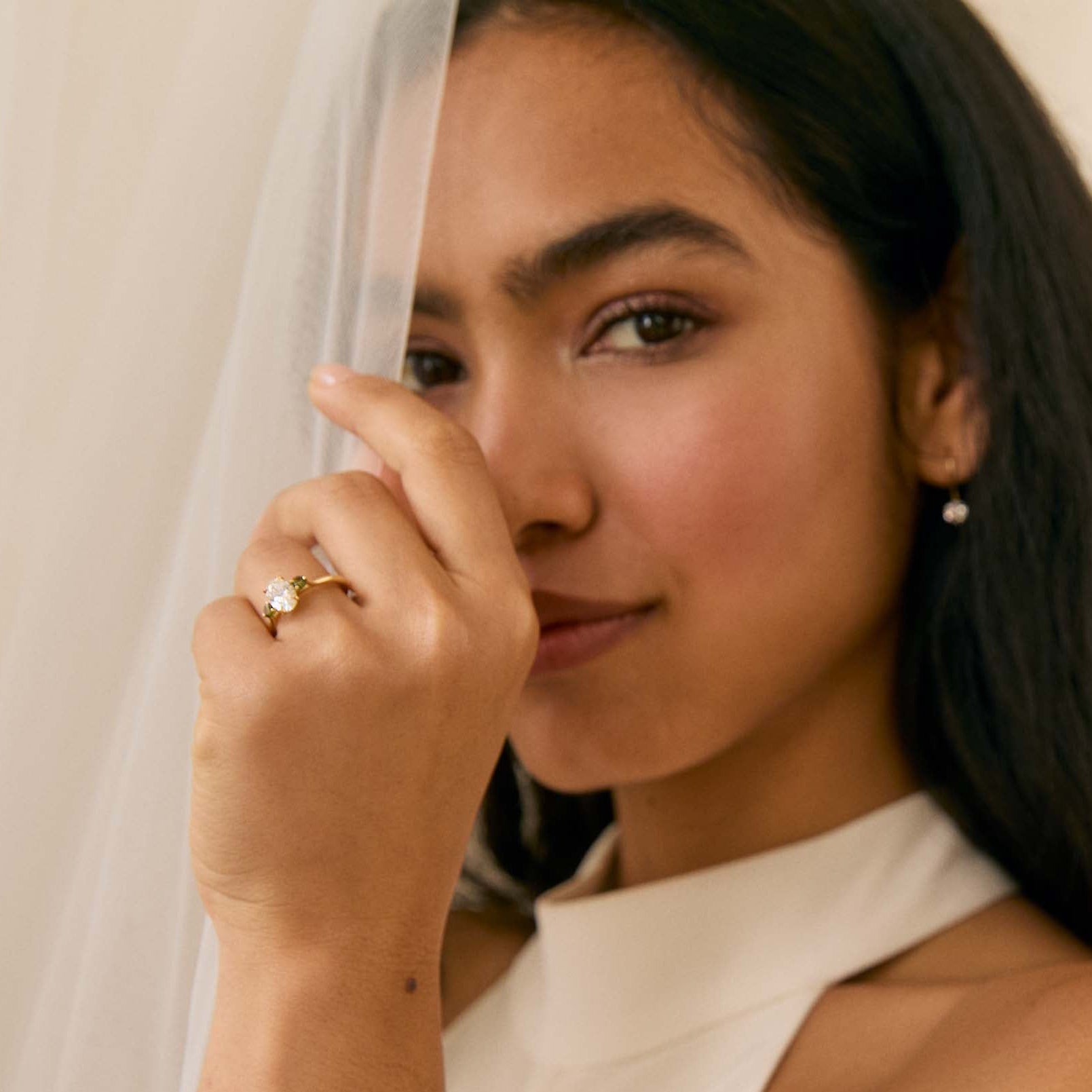 Woman wearing a white dress with a veil and a diamond engagement ring with green touramlines, posing against a plain background