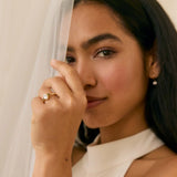 Woman wearing a white dress with a veil and a diamond engagement ring with green touramlines, posing against a plain background